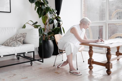 Free A woman with white hair writes in a bright, modern living room with large windows. Stock Photo