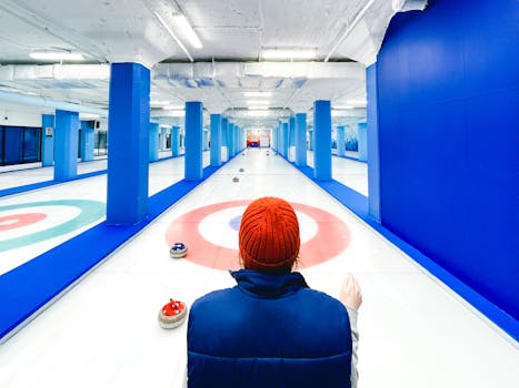 Back view of a person in a hat on an indoor curling rink, focusing on stones ahead.