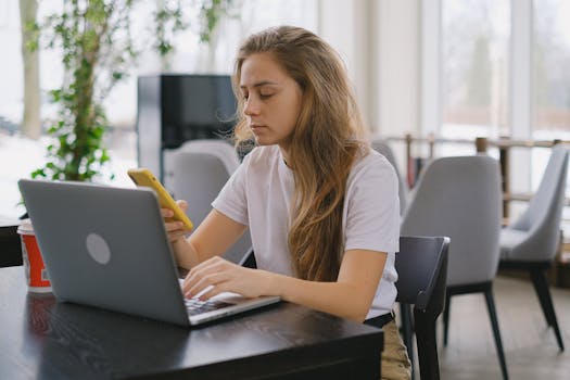 Focused woman using a laptop and smartphone at a cafe, showcasing modern multitasking.