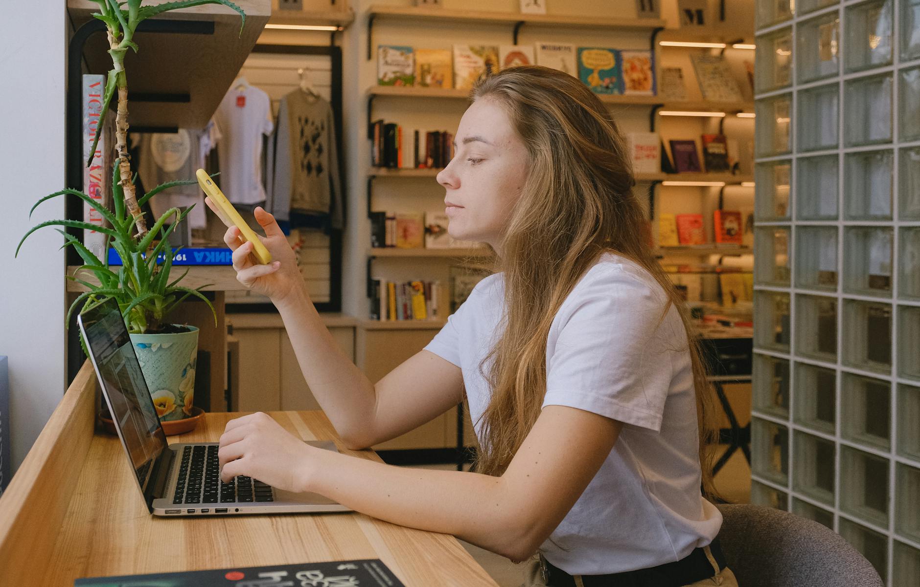 Woman multitasking with laptop and smartphone in a cozy bookstore.