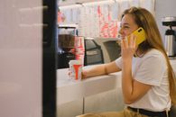 Woman in White Crew Neck Shirt Holding a Cellphone and Disposable Cup