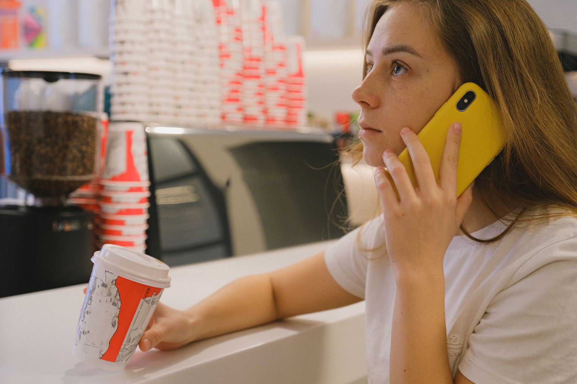 Woman with a yellow phone and coffee cup at a coffee shop counter, engaged in conversation.