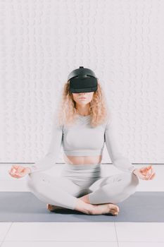 A woman in activewear using VR goggles for meditation on a yoga mat indoors.
