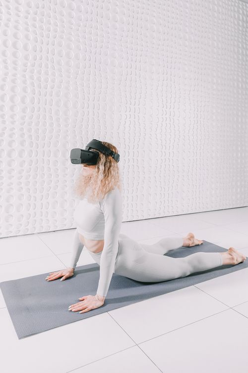 Free Woman practicing upward facing dog yoga pose with VR goggles indoors on a mat in a minimalist setting. Stock Photo