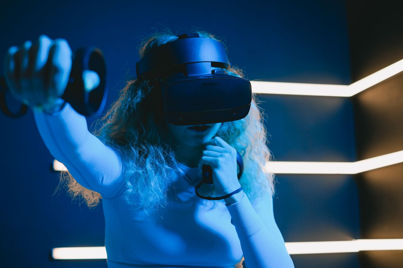 A woman immersed in virtual reality using a VR headset and controllers under blue neon lights.