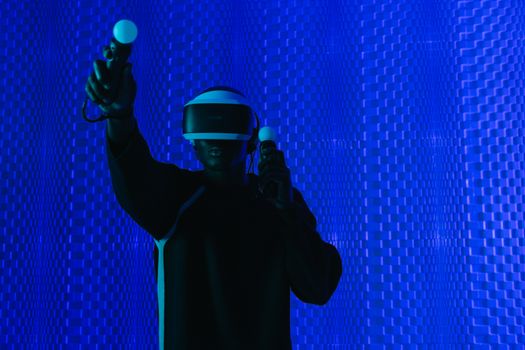 A man immersed in a virtual reality game, wearing a VR headset and controllers in a blue-lit room.