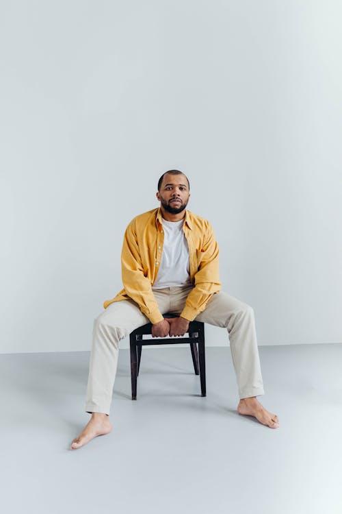 Man in Yellow Button Up Shirt and Beige Pants Sitting on Black Chair