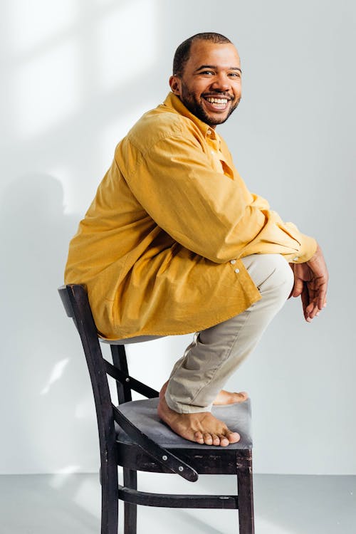 Man in Yellow Long Sleeve Shirt and Beige Pants Sitting on Black Wooden Chair Smiling