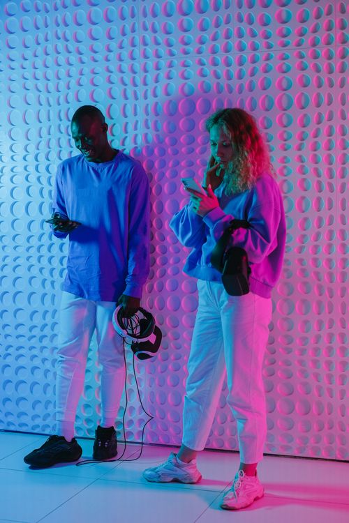 Free Young adults in a vibrant setting using mobile phones and VR headset, bathed in colorful lighting. Stock Photo