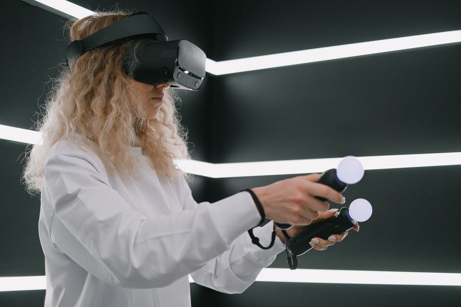 Photo by SHVETS production on Pexels A woman interacts with virtual reality using VR headset and motion controllers indoors.