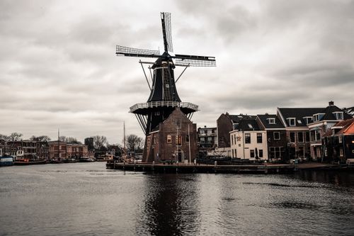 Free Scenic view of a traditional Dutch windmill by a river with urban buildings under cloudy skies. Stock Photo