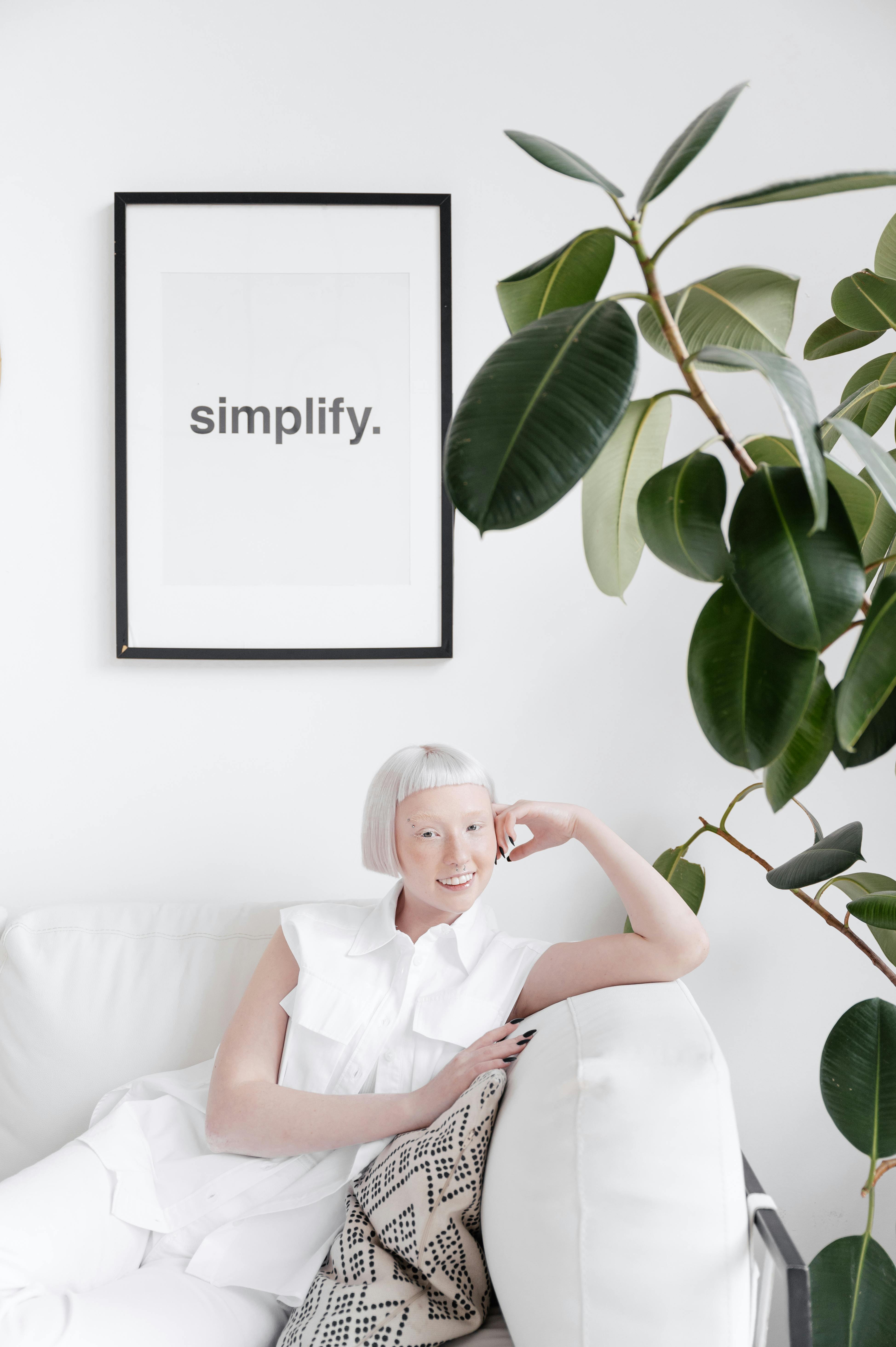 Caucasian woman sitting on a minimalist white sofa with a plant and 'simplify' poster.