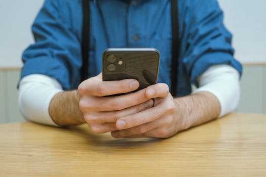 Person wearing a blue shirt holding a smartphone above a wooden tabletop indoors.
