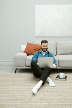 Smiling man with a beard sitting on the floor and working on a laptop in a cozy home setting.