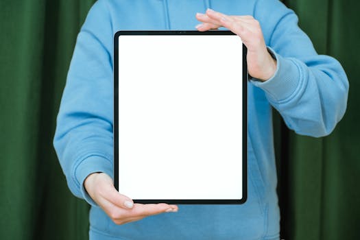 A person in a blue hoodie holding a blank tablet screen against a green curtain background.