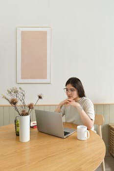 Woman sitting at a wooden table with a laptop, mug, and vase, working in a minimalistic home office.