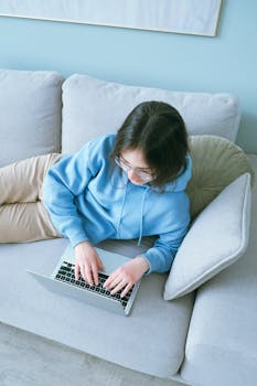 Woman wearing glasses lounging on a couch typing on a laptop, remote work scene.