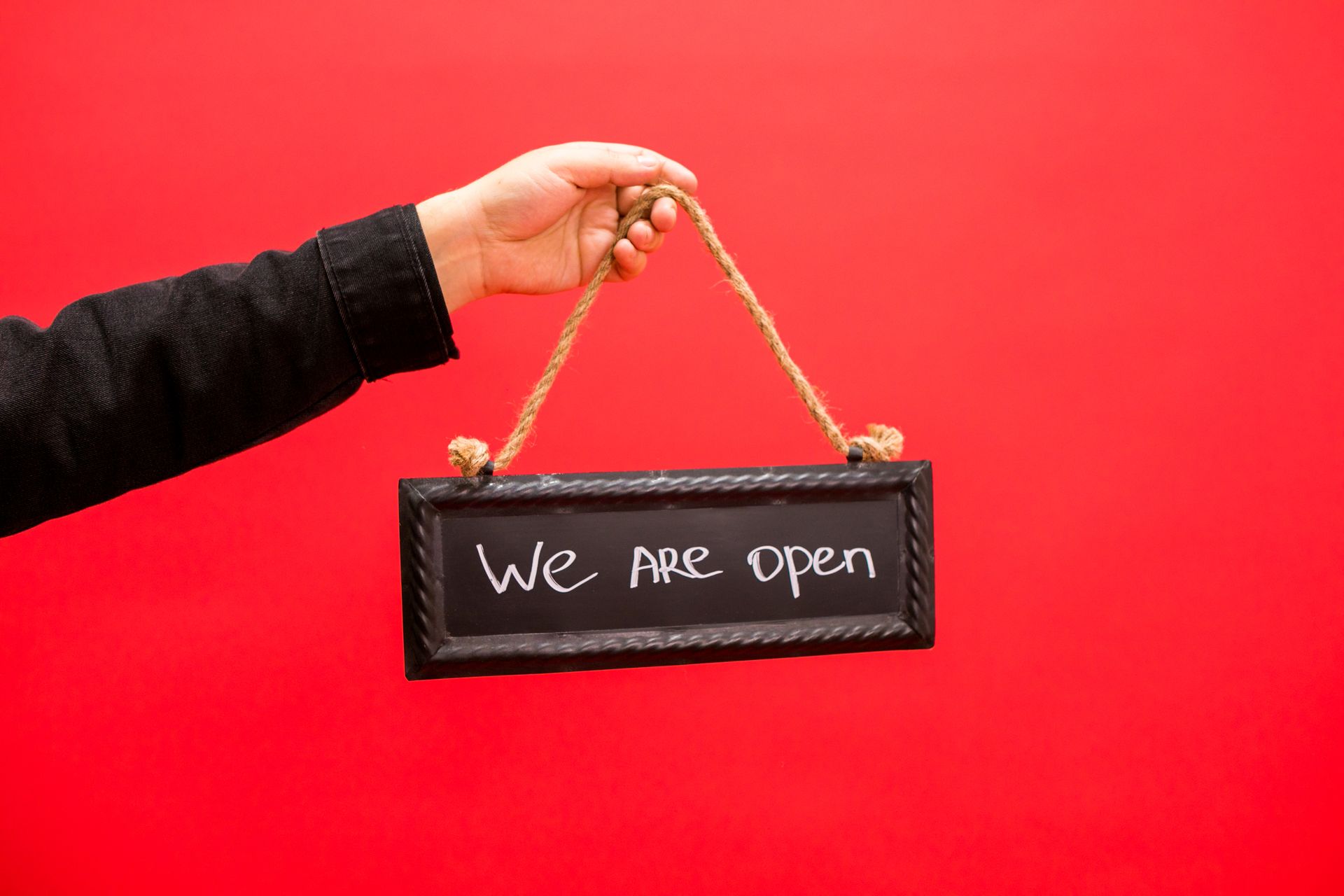 A hand holding a black sign with 'We Are Open' text, set against a vibrant red background.