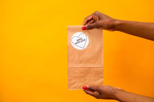 Free Hands holding a paper bag with 'Made with Love' sticker against a vibrant yellow backdrop. Stock Photo