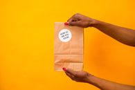 A close-up shot of hands holding a paper bag with 'Hope to See You Soon' text on a vibrant yellow background.