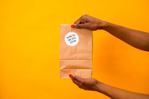 Free A close-up shot of hands holding a paper bag with 'Hope to See You Soon' text on a vibrant yellow background. Stock Photo