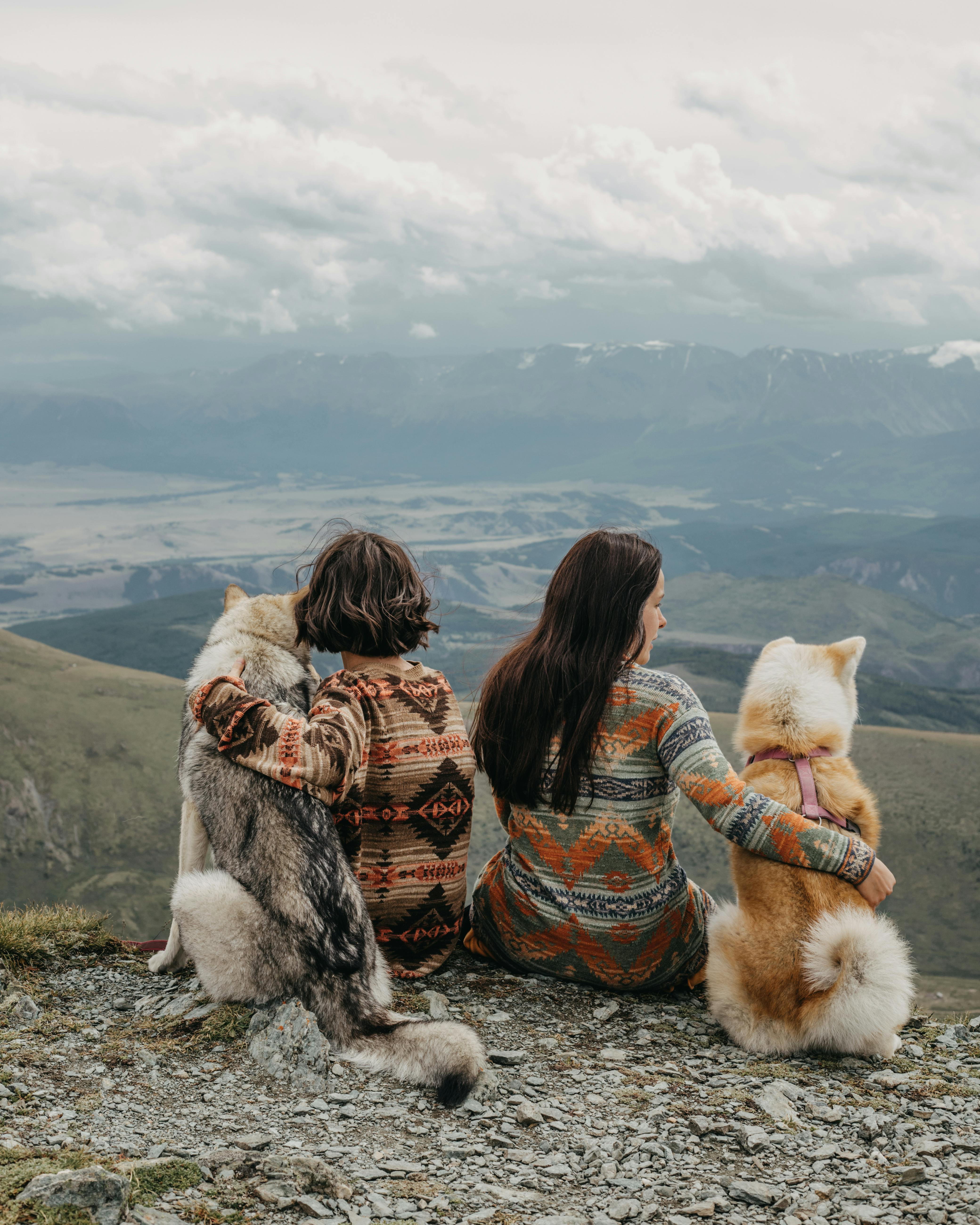 Two women and their huskies enjoy a serene mountain vista, symbolizing adventure and companionship.