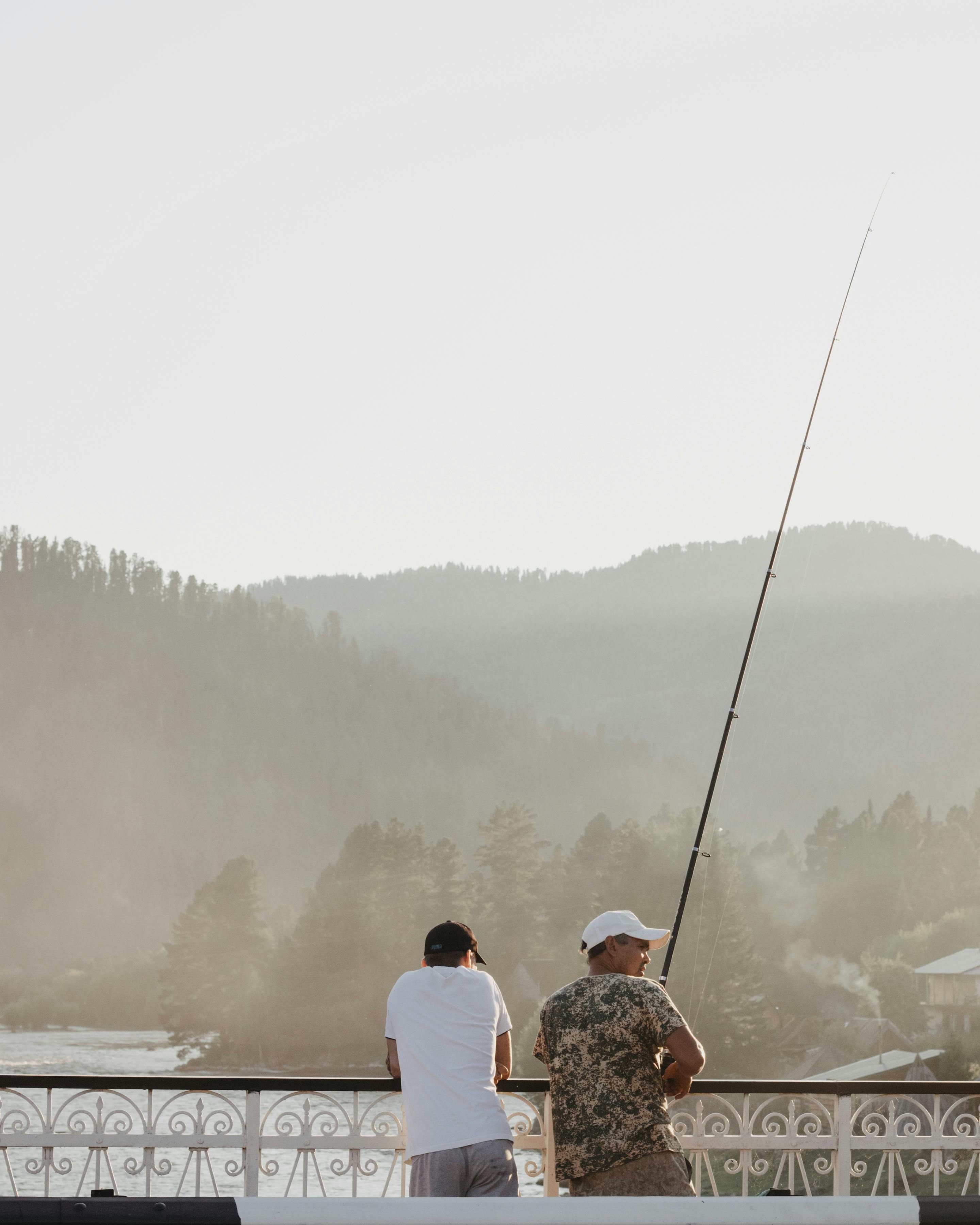 Two men fishing on a bridge with scenic mountain view.