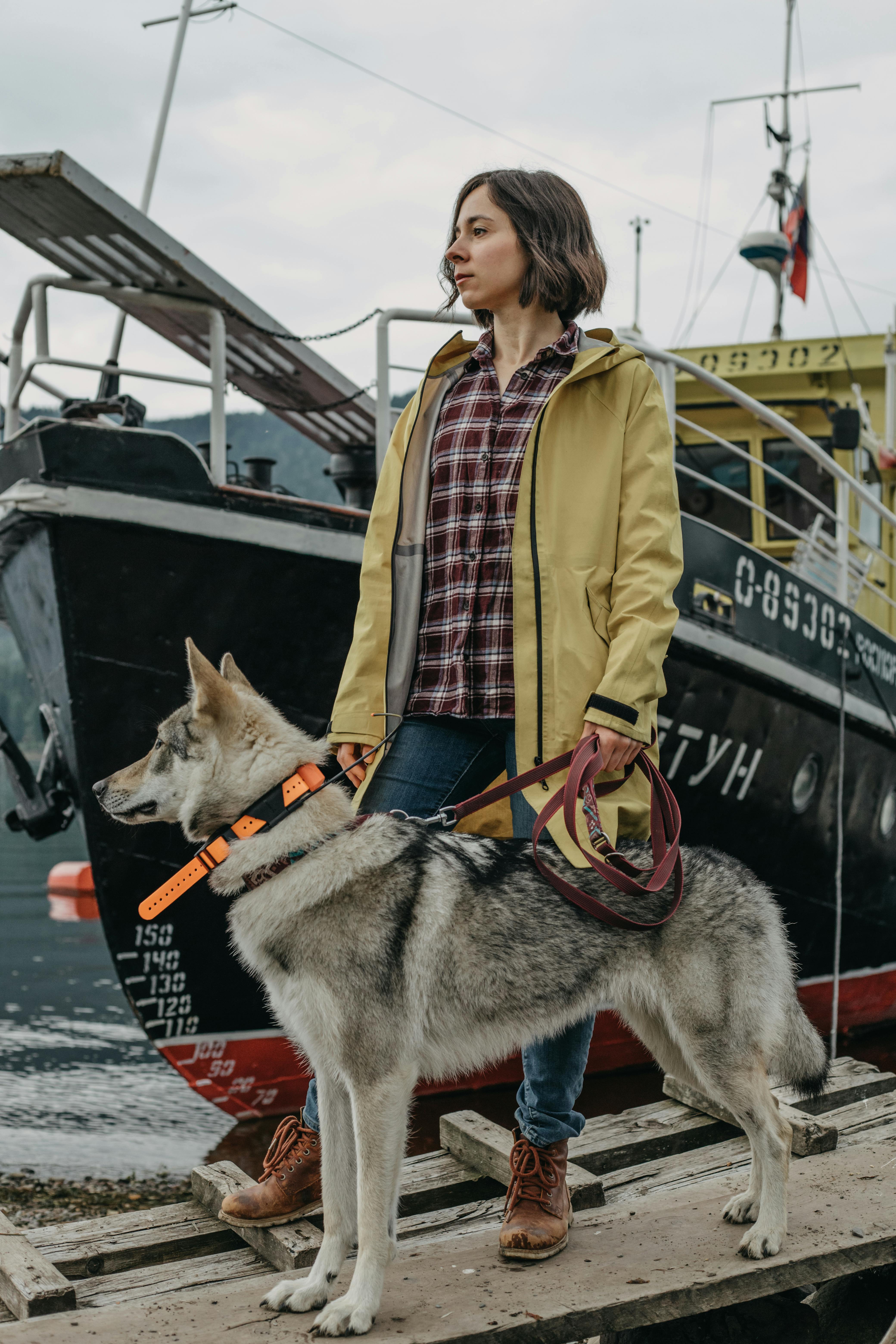 A woman in yellow jacket with a husky dog near a docked boat.