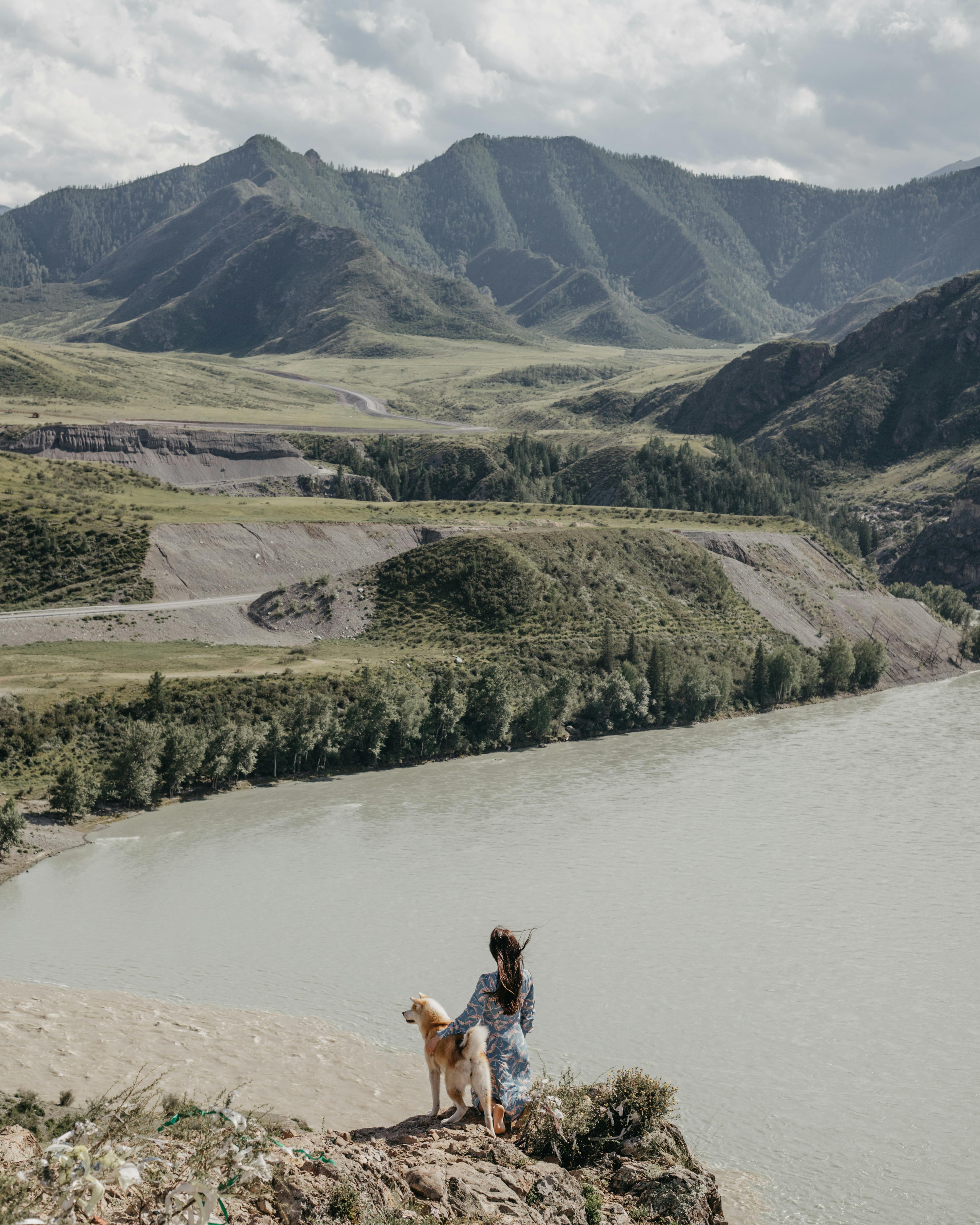 woman and her dog standing on the side of a river in mountains