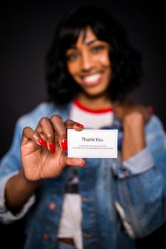 Woman with big smile holding thank you card, wearing denim jacket in a studio setting.