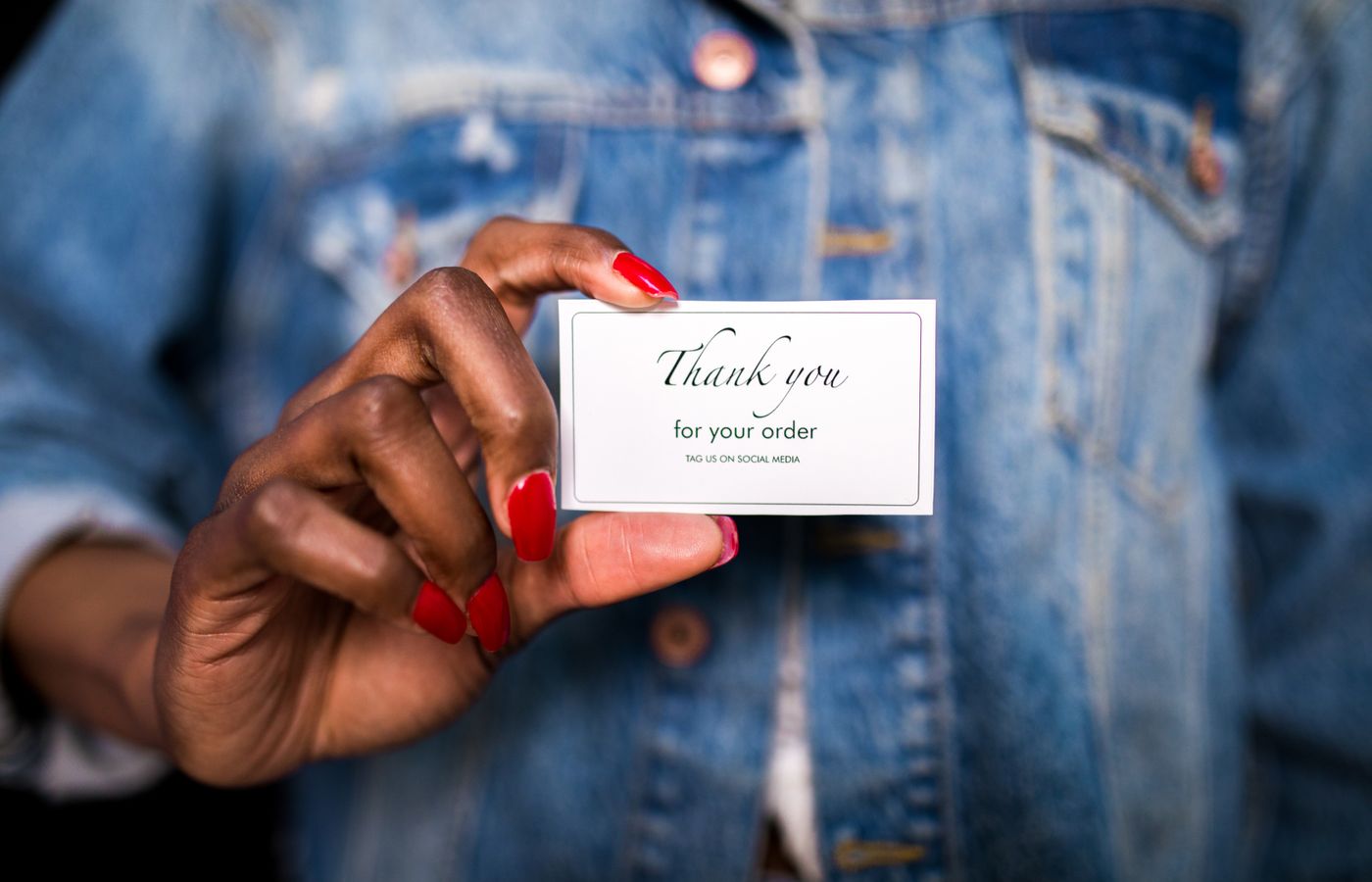 A woman in a denim jacket holds a 'Thank You' card with red nails.