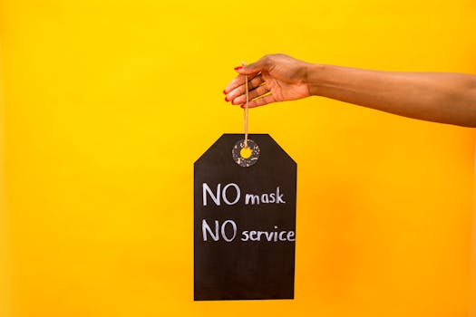 Hand holding a 'No Mask No Service' sign against a vibrant yellow background, emphasizing pandemic safety measures.