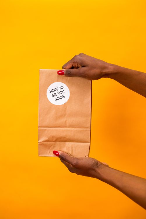 Free Photo of hands holding a paper bag with a 'Hope to See You Soon' sticker, against a vibrant orange background. Stock Photo