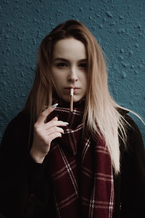 Free Young long haired blond female in scarf looking at camera with cigarette in mouth against uneven green wall Stock Photo