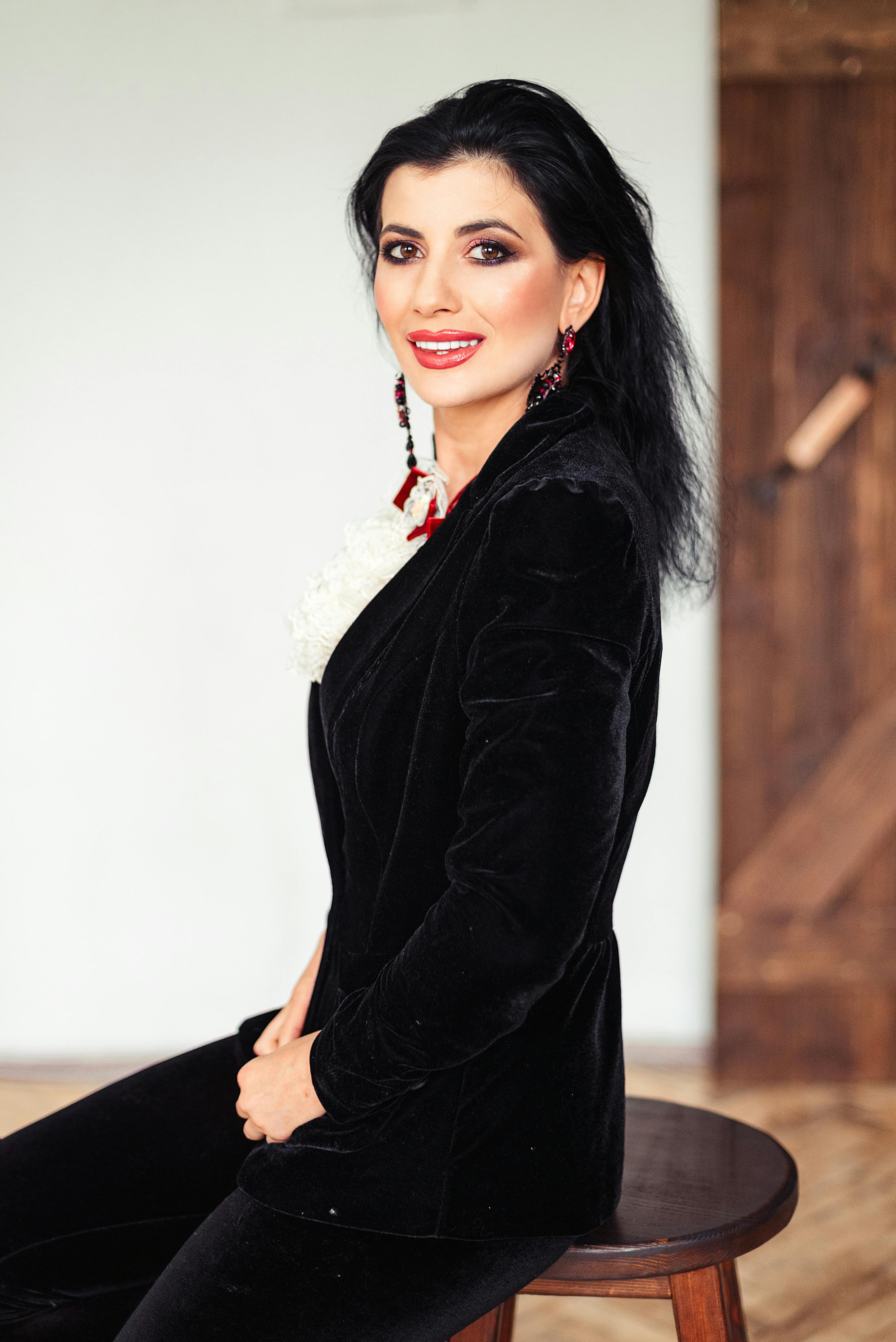 Smiling woman in a black velvet blazer sitting on a wooden stool indoors.