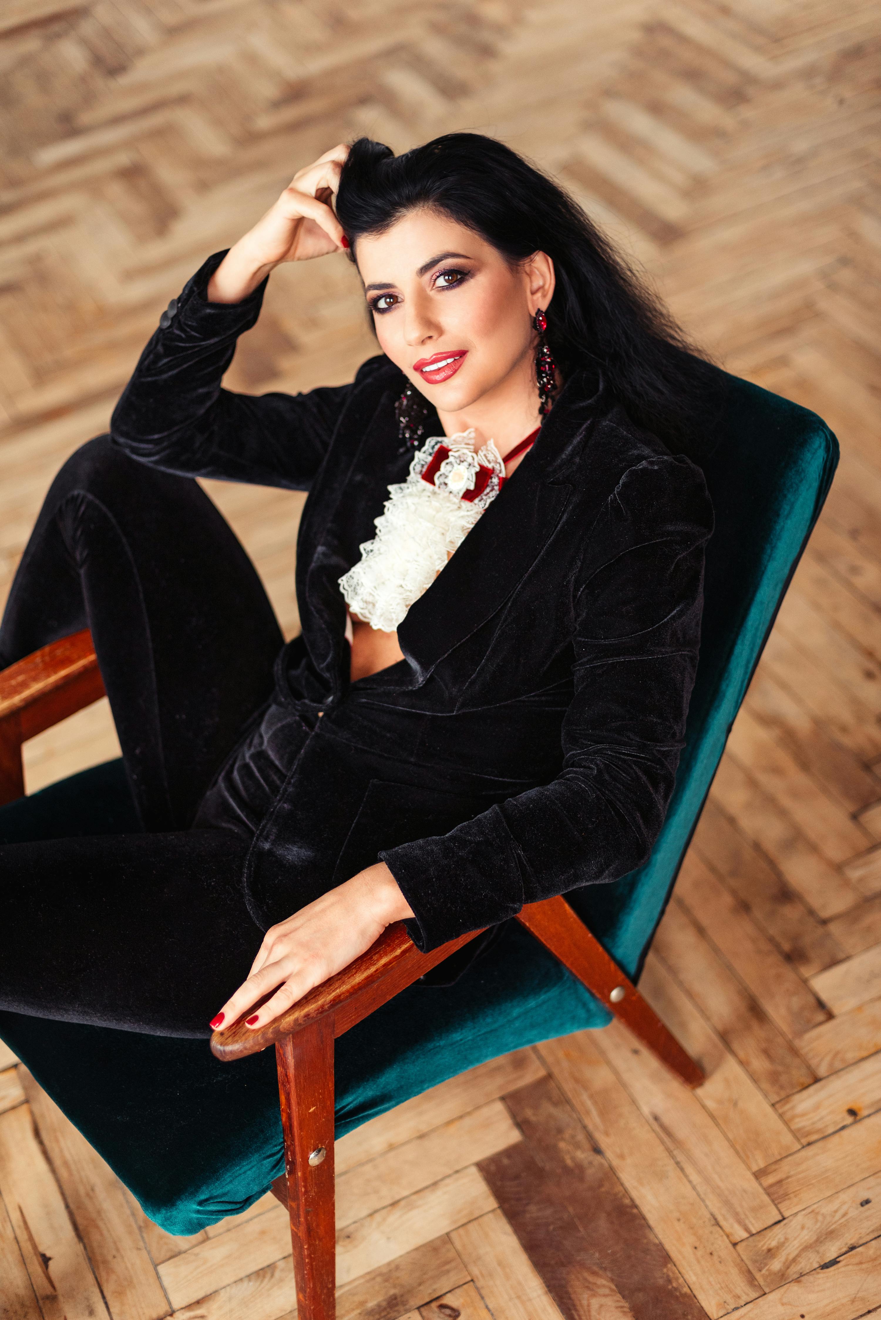 High angle of cheerful female with dark hair in stylish clothes touching hair sitting on chair in room with parquet