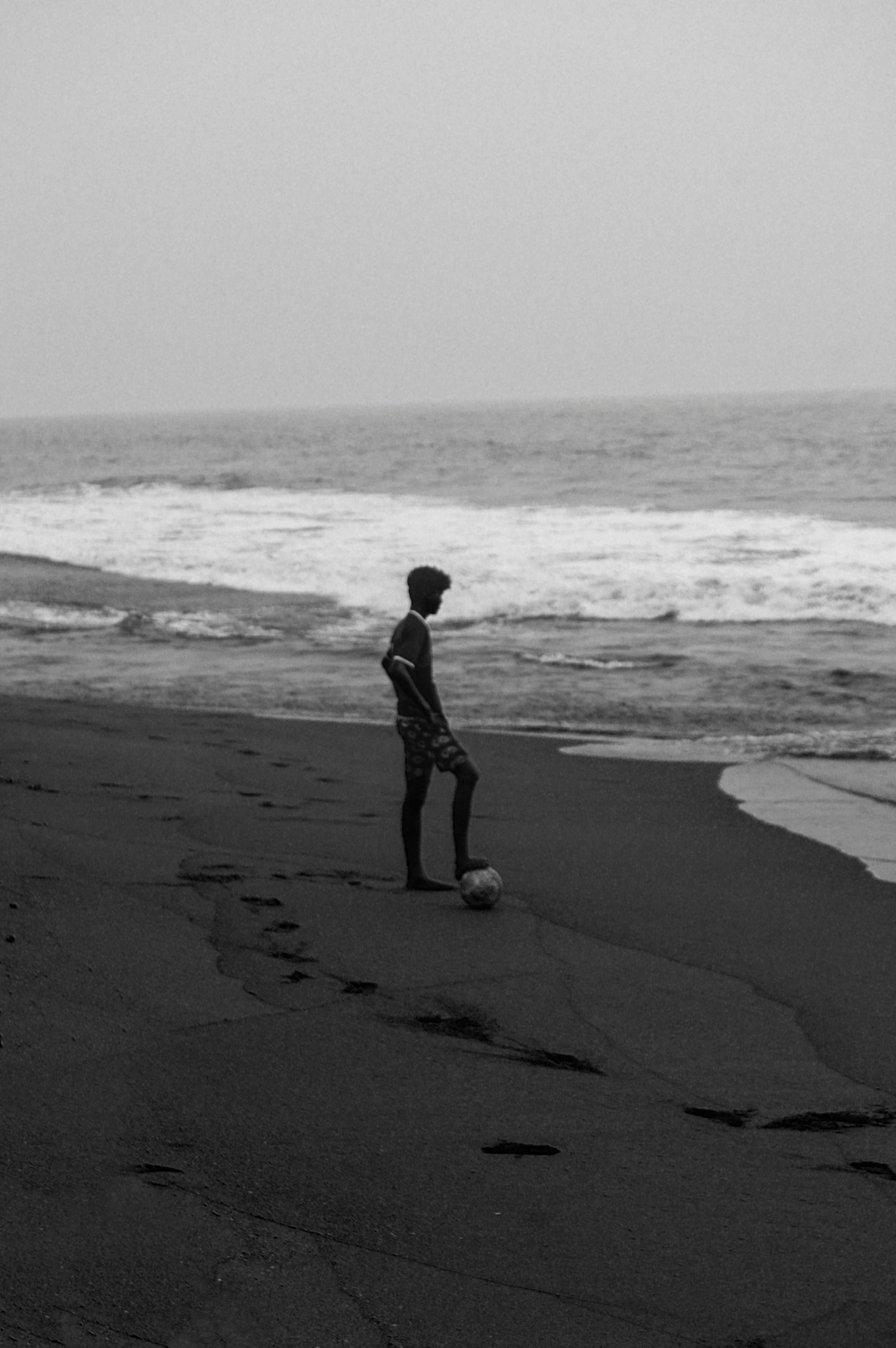 A man standing with a soccer ball on a secluded beach in Kerala, India, captured in moody monochrome.
