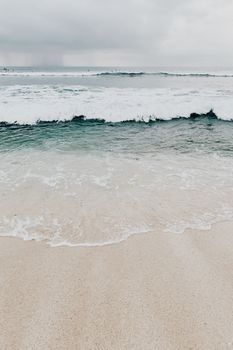 Calm ocean waves gently crash against a sandy shore under a cloudy sky.