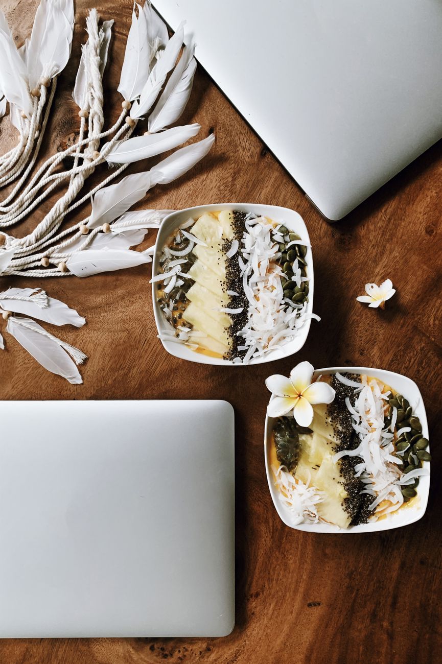 Top view of tropical smoothie bowls and laptops on wooden table, complemented by decorative feathers.
