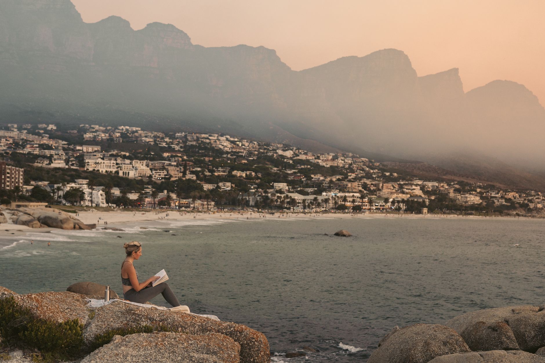 A woman enjoys reading on the rocks overlooking a scenic beach and mountains in Cape Town at sunset.