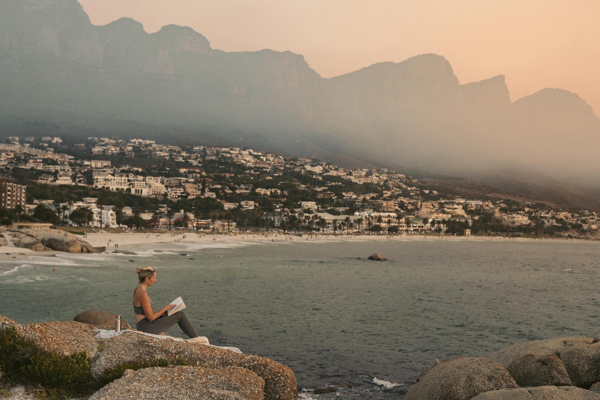 A woman enjoys reading on the rocks overlooking a scenic beach and mountains in Cape Town at sunset.