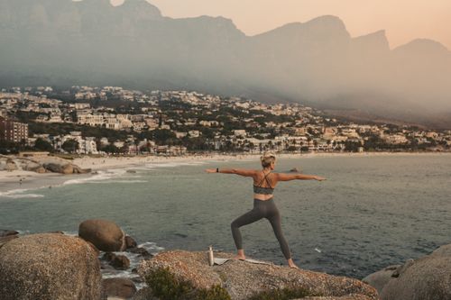 Free Woman practicing yoga warrior pose with scenic view of Cape Town coastline at sunset, promoting wellness. Stock Photo