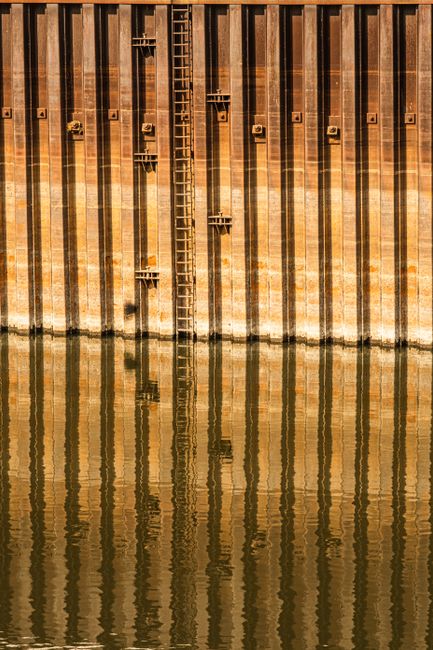 Rusty metal wall with vertical lines and ladder reflecting in water, creating a symmetric abstract pattern.
