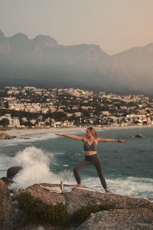 Free Woman practicing yoga warrior pose on rocks by Cape Town coastline during sunset. Stock Photo