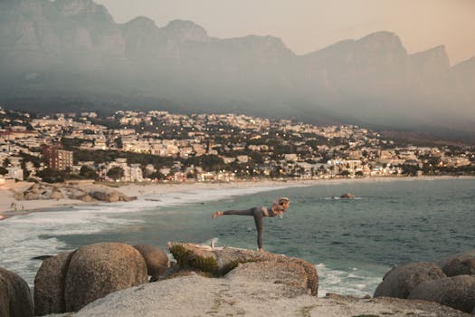 Woman practicing yoga on Cape Town's scenic coastline with mountain backdrop.
