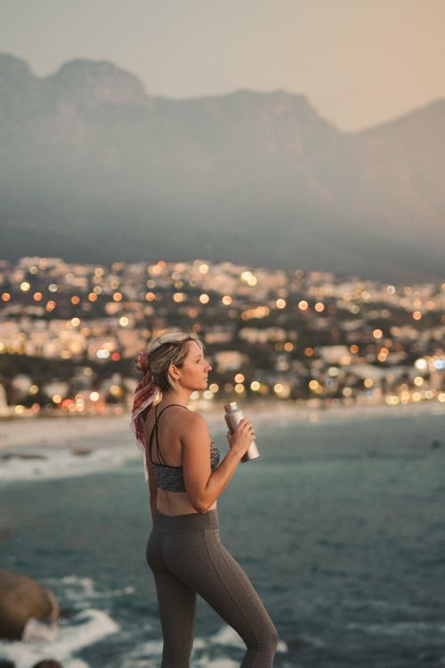 Free A woman stands by the ocean with a view of Cape Town city lights and mountains at dusk, exuding tranquility and adventure. Stock Photo