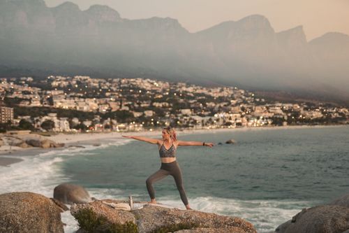 Free A woman performs the warrior pose on rocks overlooking the ocean at sunset in Cape Town, showcasing tranquility. Stock Photo