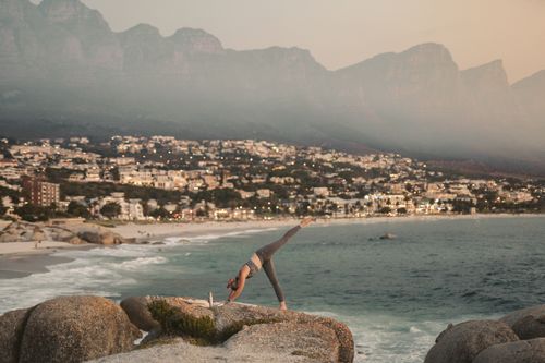 Free A woman practices yoga on a cliff overlooking a beach in Cape Town with mountains in the background. Stock Photo