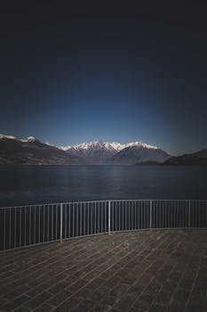 Waterfront with fence near calm river flowing near mountain ridge covered with hoarfrost on winter day in nature against blue sky