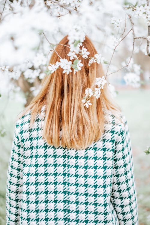 Back view of unrecognizable female with long hair standing under branches of tree with blooming fragrant flowers